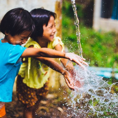 Two children smile as they run their hands under water running from a spigot.